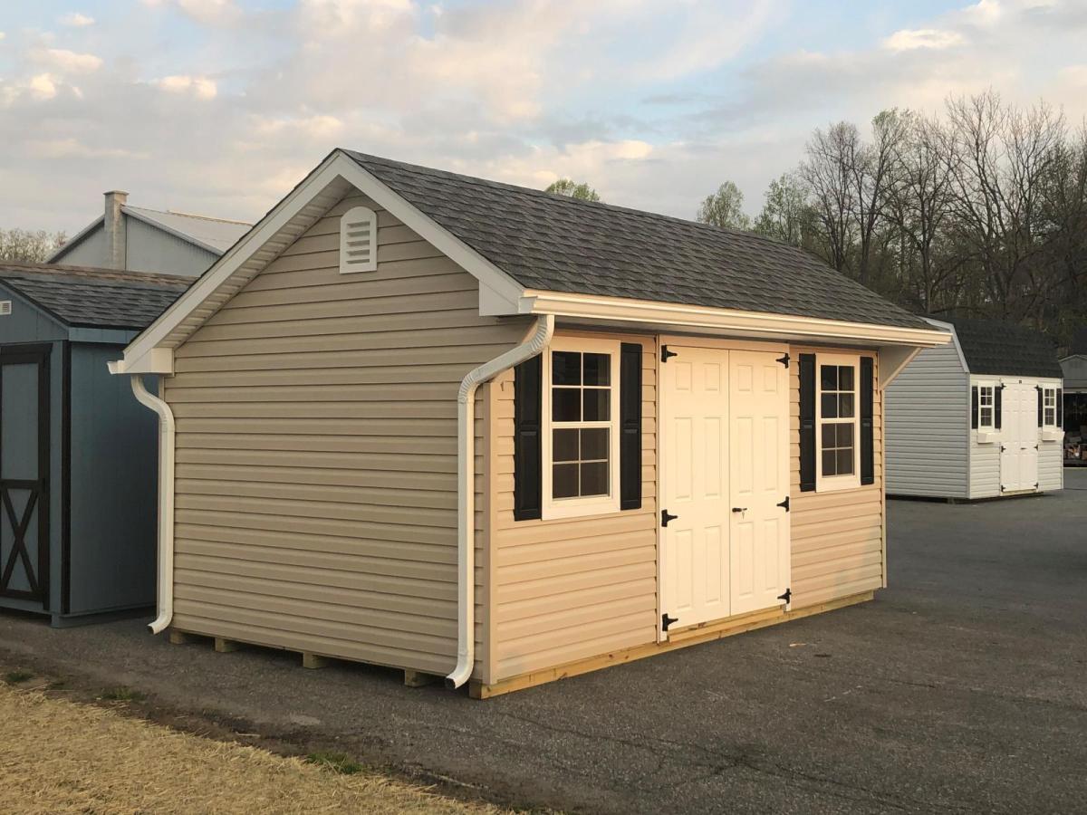 10x16 Classic Quaker Shed with tan siding, white rain spouts, a set of white double doors, white windows with black shutters, and a dark gray asphalt roof.