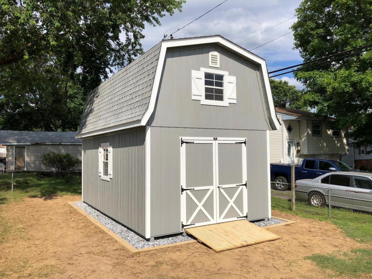 Elite Classic Garden Dutch Barn garage with 2 floors, gray wood siding, white trim, double doors, 2 white windows with white shutters, and a matching gray asphalt roof on a stone foundation in a yard.