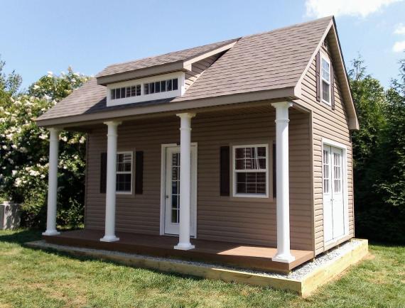 Elite Classic Garden Cottage with white pillars, a covered front porch, tan siding, light brown asphalt roofing, a white transom window, a white door with glass panels, and white windows with black shutters.