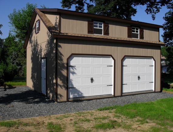 2 story, 2 car garage with tan brown siding, brown trim, brown shutters, 2 white garage doors, a single entry side door, and brown roofing.