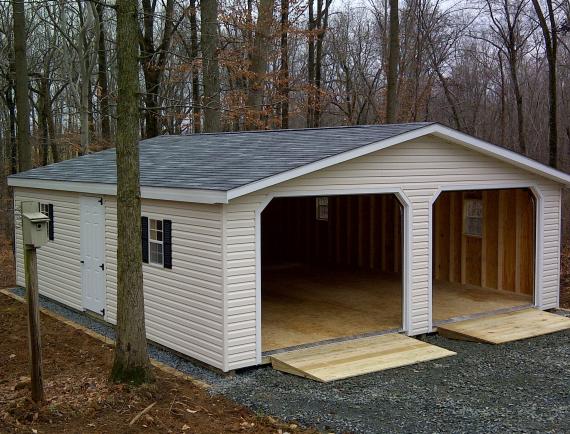 Doublewide Garage with beige siding, gray asphalt roofing, 2 open garage doors with ramps, and white windows with black shutters.