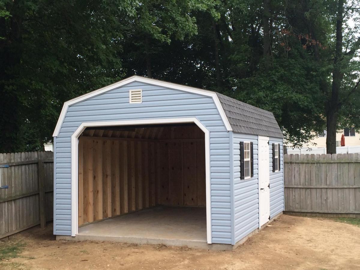 Dutch Barn Garage with light blue vinyl siding, white trim, an open garage door, a white single-entry side door, 2 white windows with black shutters, and a gray asphalt roof in front of a wood fence in a backyard.