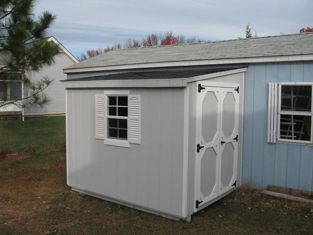 Lean-To Shed with wood siding painted light gray, white trim, a white window with white shutters, double doors, and a black asphalt roof against a light blue building.