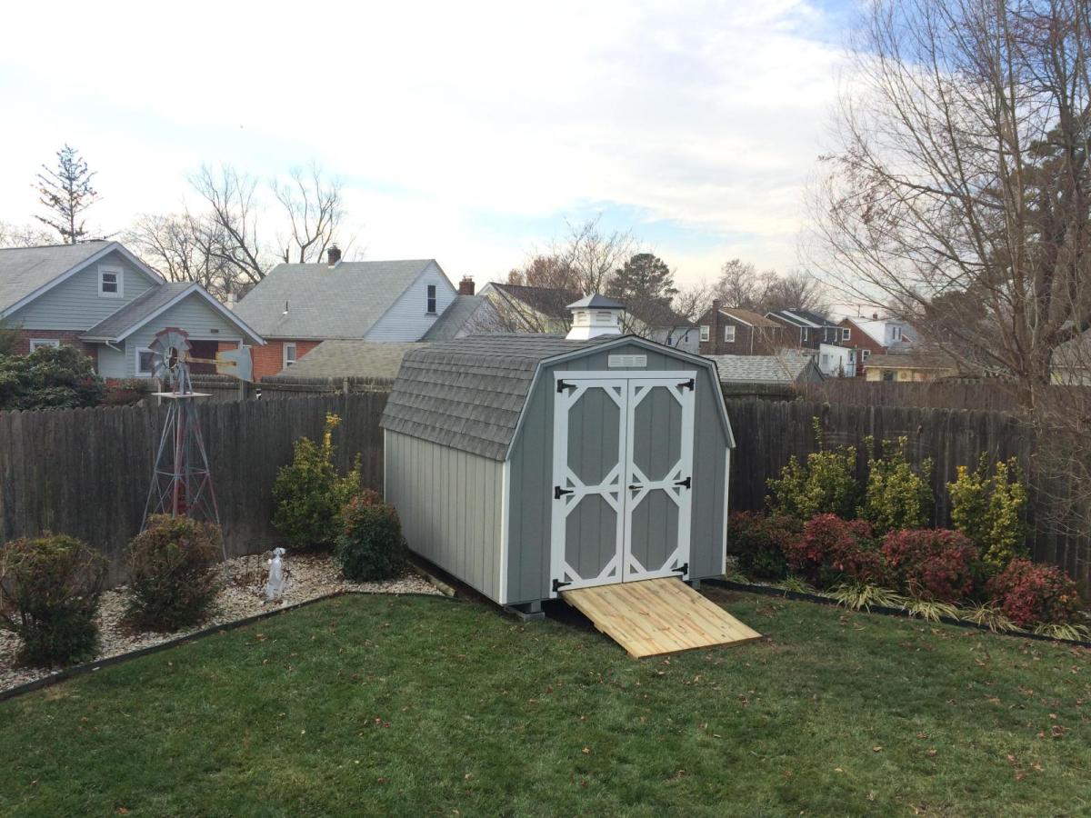 Mini Barn shed with gray wood siding, white trim, double doors, a ramp, and a gray asphalt roof sitting the the corner of a backyard in front of a brown wood fence.