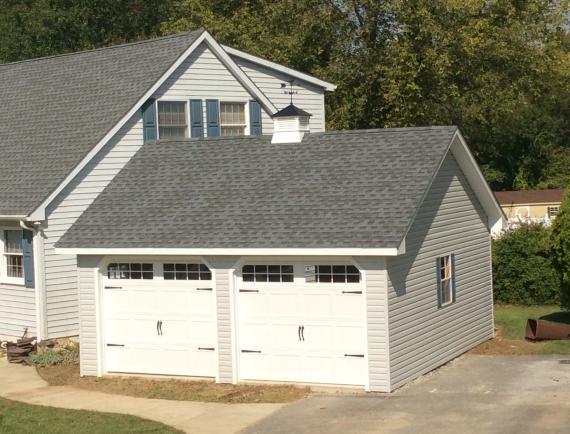 2 car garage with 2 white garage doors with windows, beige vinyl siding, white trim, 1 white window with gray shutters, a gray asphalt roof, and a white cupola.